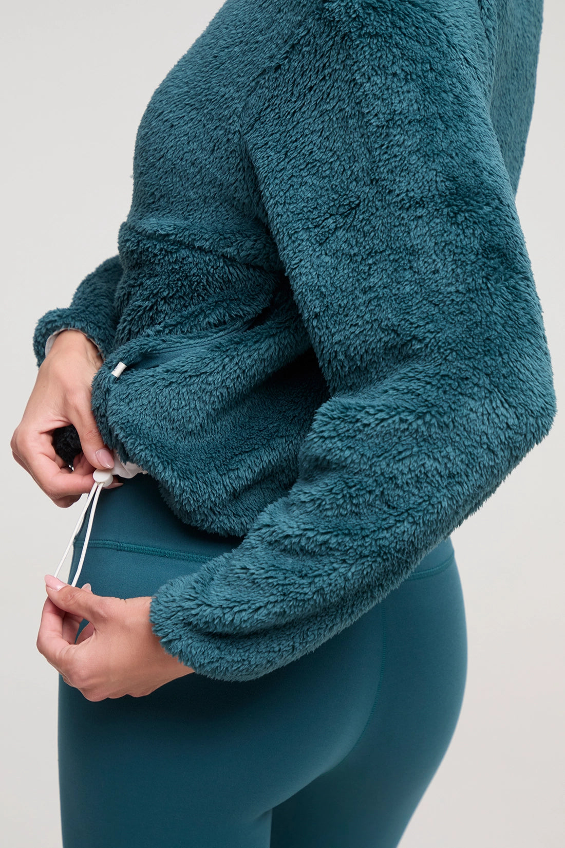 Close-up of a woman wearing a teal fuzzy fleece long-sleeve top and matching teal leggings, adjusting the drawstring at the back of the top against a light gray studio background.