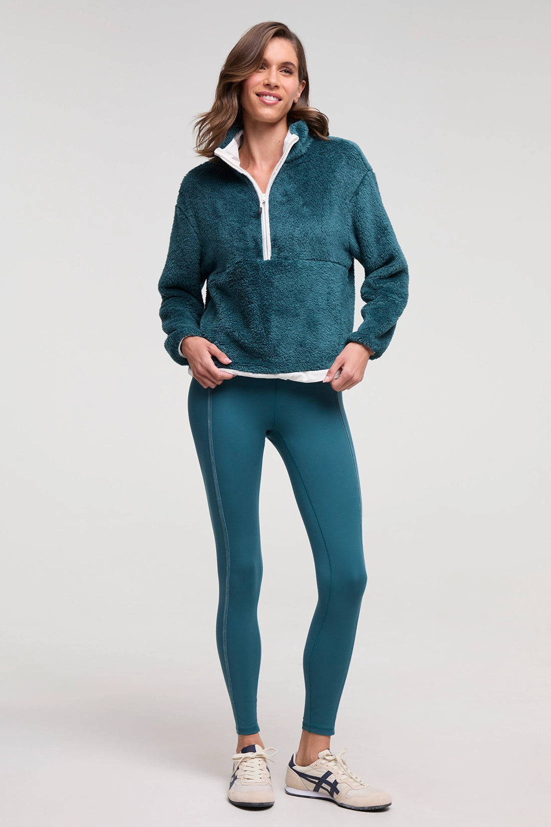 A woman modeling a teal fleece quarter-zip pullover and matching teal leggings with white sneakers, photographed against a light gray studio background. 