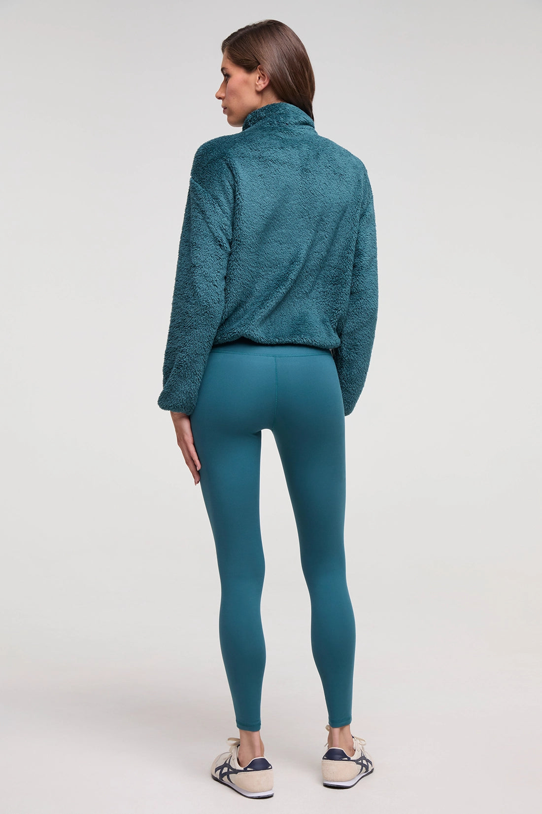 A woman is shown from the back wearing a textured teal fleece pullover and matching teal leggings, paired with white sneakers, against a plain light gray studio background. 