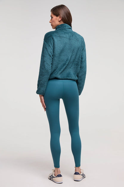 A woman is shown from the back wearing a textured teal fleece pullover and matching teal leggings, paired with white sneakers, against a plain light gray studio background. 