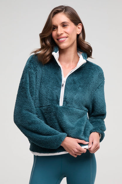 A woman wearing a teal fleece quarter-zip pullover with a white inner collar and white hem, paired with matching teal leggings, against a light gray studio background. 