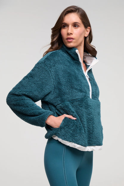 A woman wearing a teal fleece quarter-zip pullover with a white inner collar and hem, paired with matching teal leggings, against a light gray studio background. 