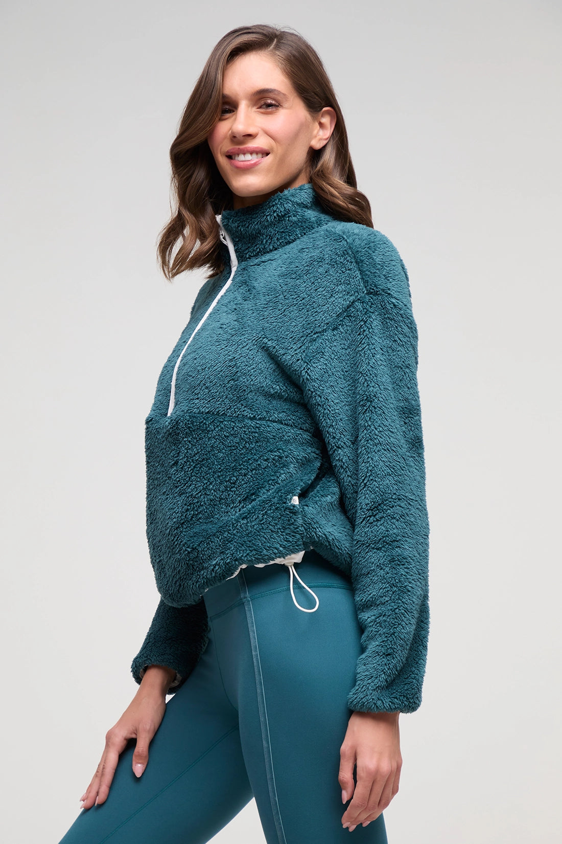 A woman wearing a teal fleece quarter-zip pullover and matching teal leggings, photographed against a light gray studio background. 